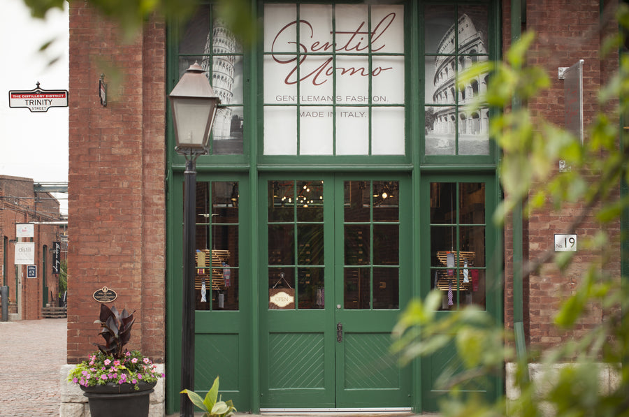 Green double doors with 'Gentil Uomo' sign above, surrounded by brick building and plants.