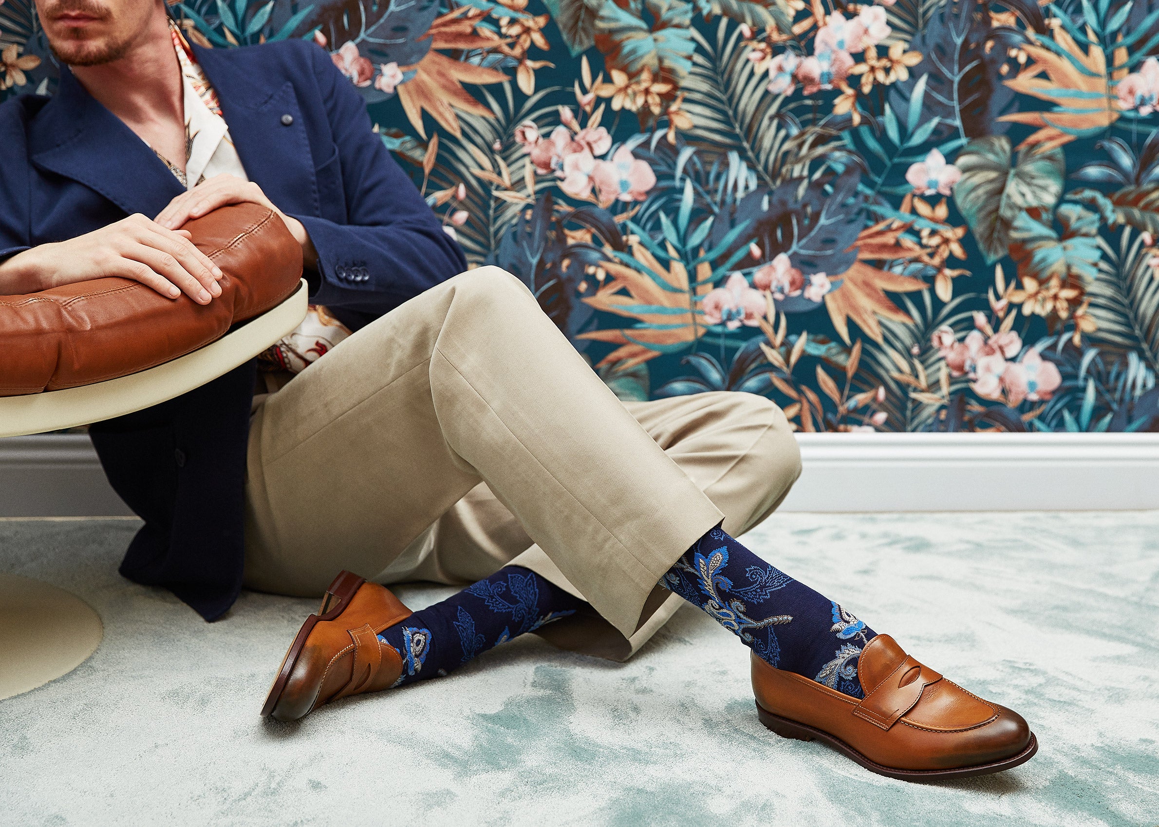 Man sitting on a rug wearing brown shoes and patterned socks with a floral wallpaper background.